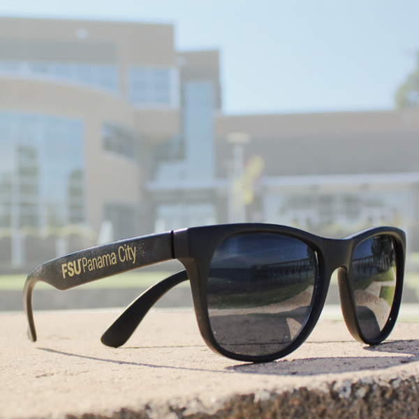 Close up of FSU Panama City branded sunglasses with the back of the Holley Academic Center faded out in the background